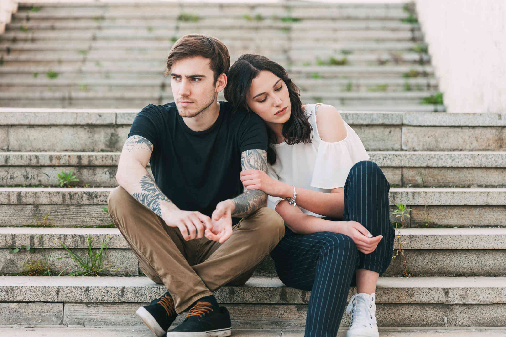 A couple is sitting outdoors, the woman leans her head on her partner’s shoulder while holding his arm, as the man looks away, deep in thought.